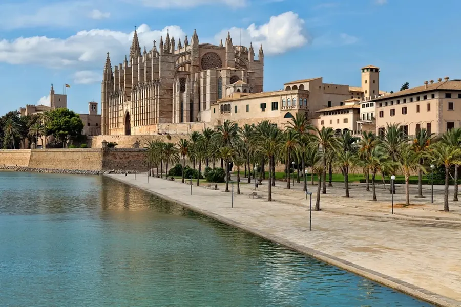 Die Kathedrale La Seu in Palma über dem Wasserbecken des Parc de la Mar, davor Palmen und eine breite Promenade