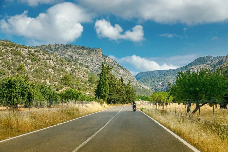 Ein Motorradfahrer auf einer leeren Landstraße zwischen Mandelbäumen und Zypressen, dahinter die Berge der Serra de Tramuntana